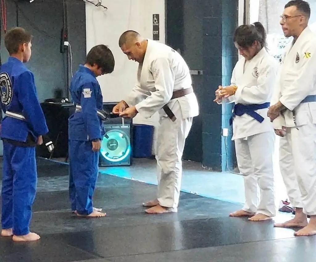 A coach ties a new belt on a young student during a belt promotion, while other students wait their turn.